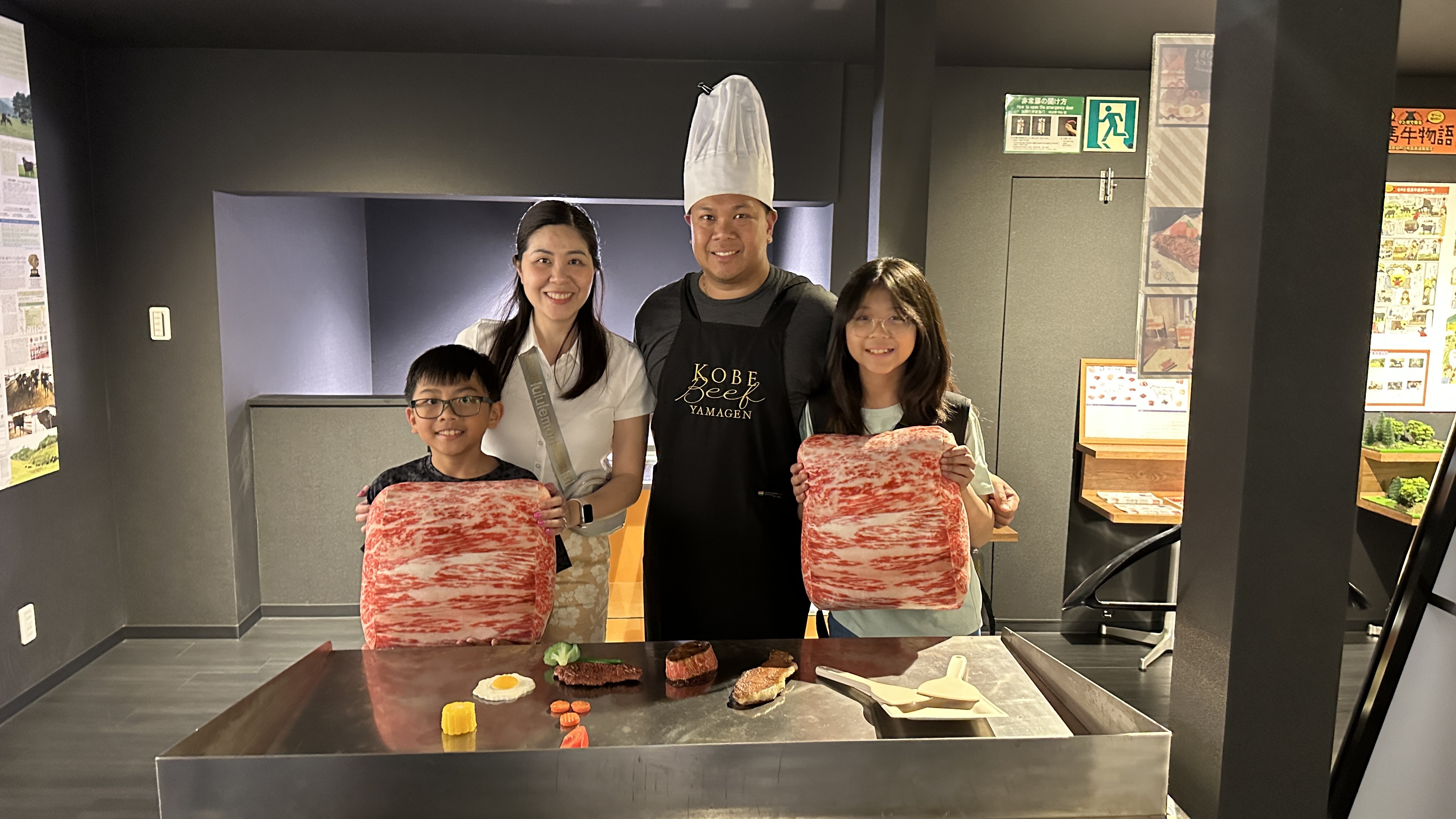A smiling family of four poses with a chef at a teppanyaki grill station. The chef wears a tall white hat and a black apron that reads "Kobe Beef Yamagen." The two children hold large, realistic cushions shaped like marbled slices of wagyu beef. In front of them on the grill are realistic food props, including steak, vegetables, and a fried egg. The setting is a sleek, modern interior, likely part of a culinary or Kobe beef experience.