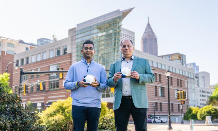 1.	Georgia Tech Professor Emmanouil “Manos” Tentzeris and Ph.D. student Marvin Joshi hold a lens enabled backscatter system device in front of a midtown Atlanta skyline. 