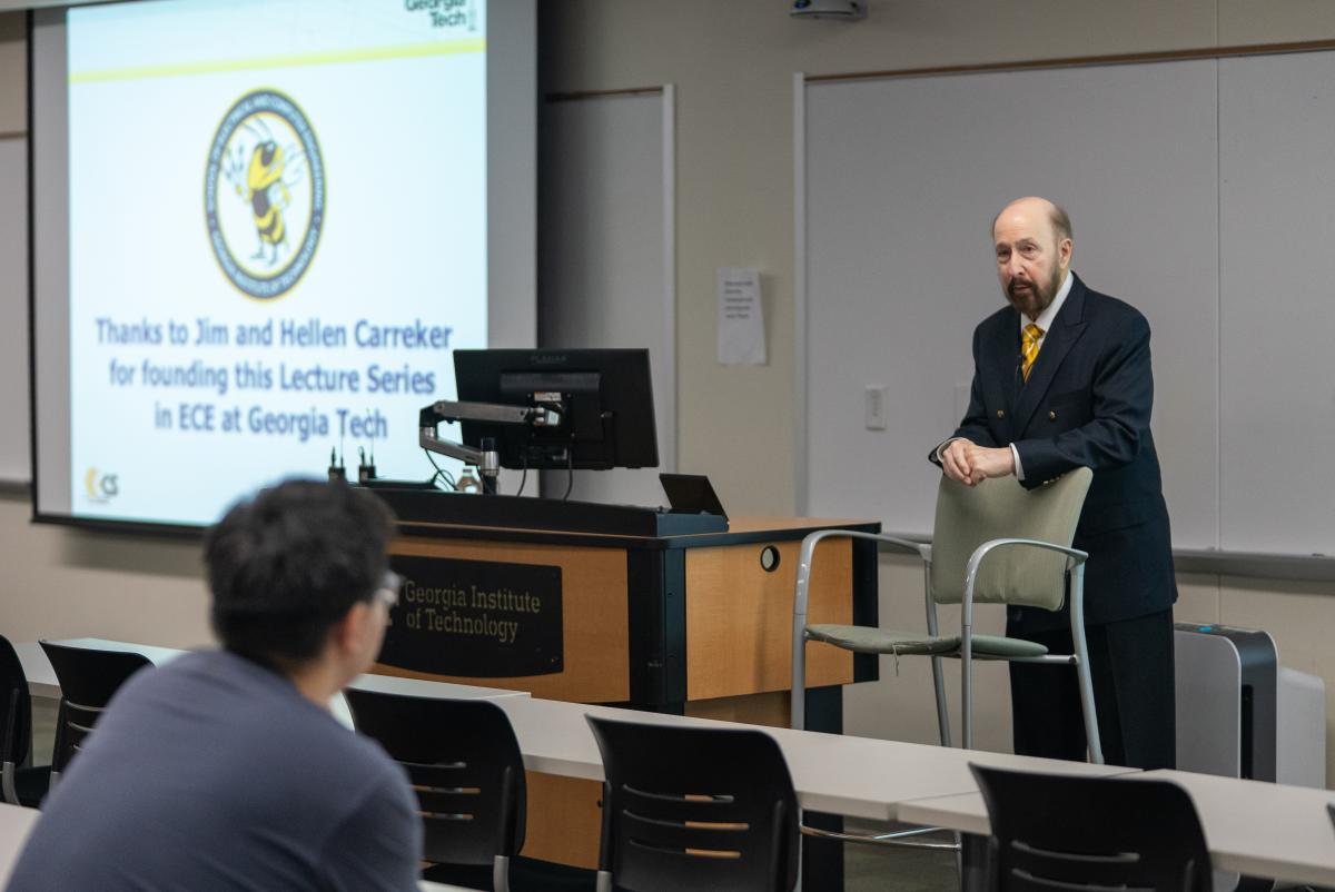 Professor Russell Dupuis stands beside a chair at the front of the room while answering audience questions following his lecture.