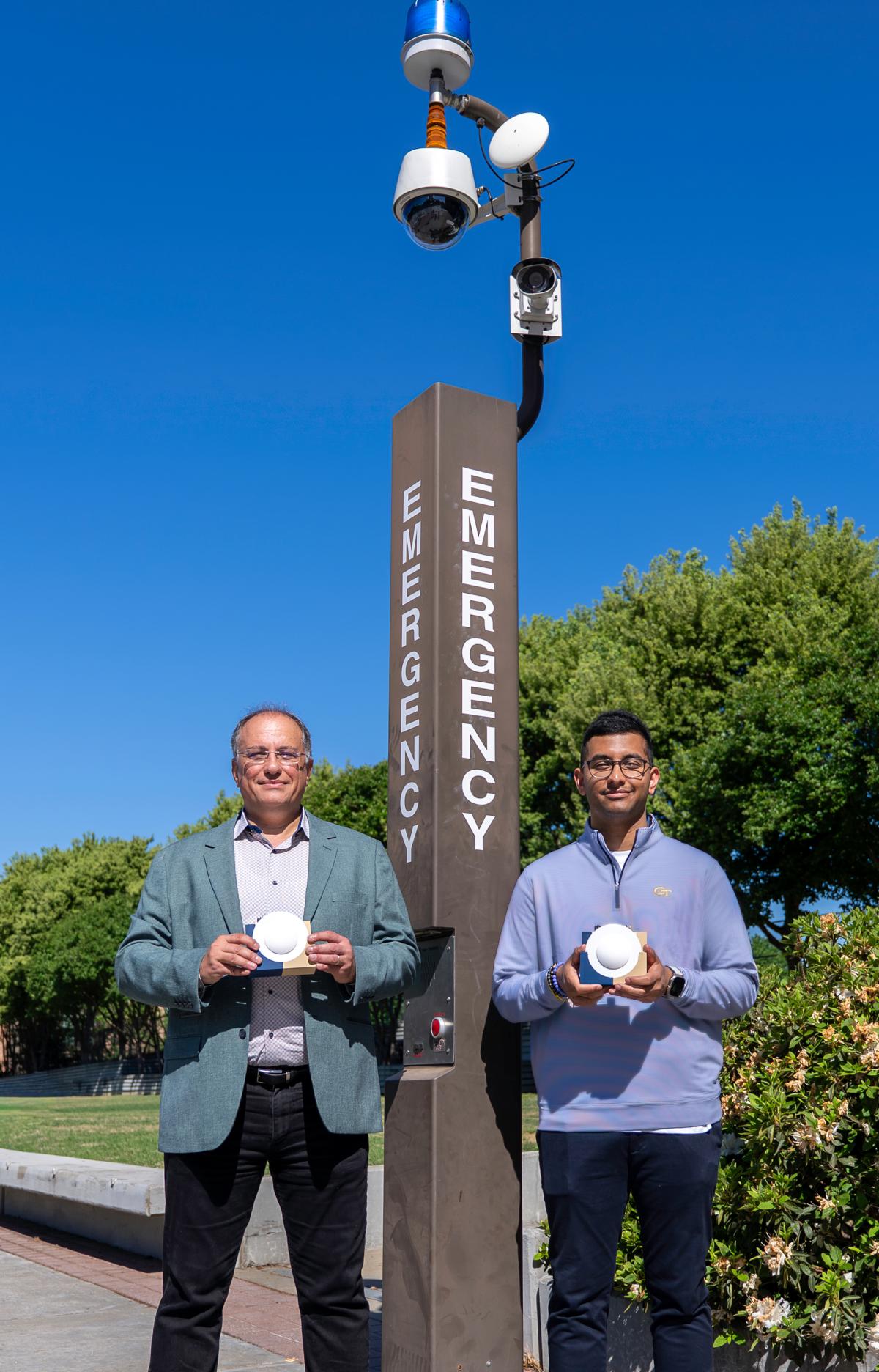 Georgia Tech Professor Emmanouil “Manos” Tentzeris and Ph.D. student Marvin Joshi hold a lens enabled backscatter system near existing campus emergency infrastructure