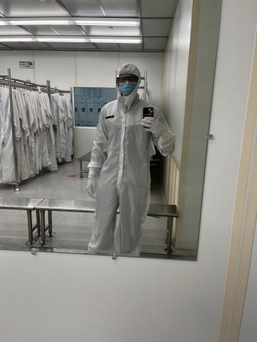Calib Lanier wearing a full cleanroom suit, face mask, safety glasses, and gloves takes a mirror selfie in a gowning area lined with cleanroom garments and lockers.